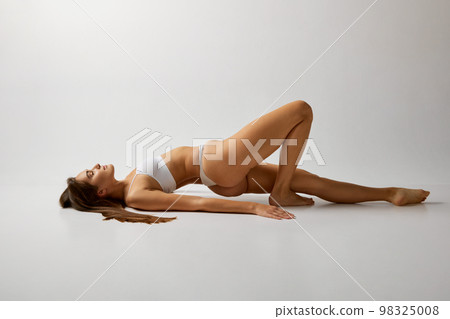 Femininity. Slim body. Portrait of young beautiful girl posing in white underwear over grey studio background. Concept of body and skin care, fitness 98325008