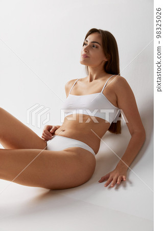 Femininity. Portrait of young beautiful girl posing in white underwear over grey studio background. Concept of body and skin care, fitness Femininity. Portrait of young beautiful girl posing in white underwear over grey studio background. Concept of body and skin care, fitness 98325026