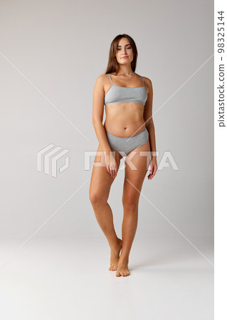 Self-care. Portrait of young beautiful girl with slim body posing in cotton underwear over grey studio background. Concept of body and skin care, fitness Self-care. Portrait of young beautiful girl with slim body posing in cotton underwear over grey studio background. Concept of body and skin care, fitness 98325144