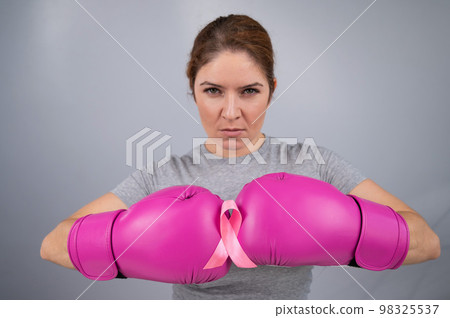 Caucasian woman in pink boxing gloves with a pink ribbon on her chest on a gray background. Fight against breast cancer.  98325537