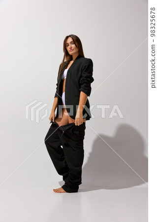 Getting ready. Portrait of young beautiful girl posing in underwear and black classic suit over grey studio background. Concept of body and skin care, fitness 98325678