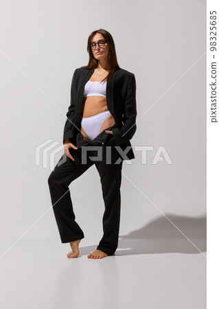 Business lady. Portrait of young beautiful girl posing in underwear and black classic suit over grey studio background. Concept of body and skin care, fitness Business lady. Portrait of young beautiful girl posing in underwear and black classic suit over grey studio background. Concept of body and skin care, fitness 98325685