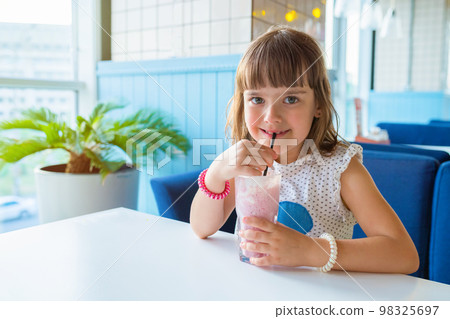 Little girl sitting at a table in a restaurant, with a milkshake in her hands. Healthy nutrition and lifestyle Little girl sitting at a table in a restaurant, with a milkshake in her hands. Healthy nutrition and lifestyle 98325697
