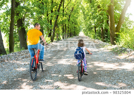 A happy dad with daughter ride bikes in the park. Back view A happy dad with daughter ride bikes in the park. Back view 98325804