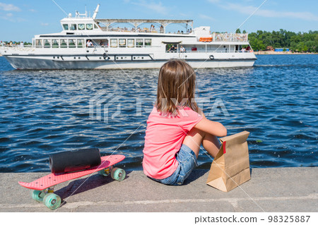 A child is sitting on a river bank with a skateboard and music speaker, a bag with products at the river and ship background. An active lifestyle. A back view 98325887