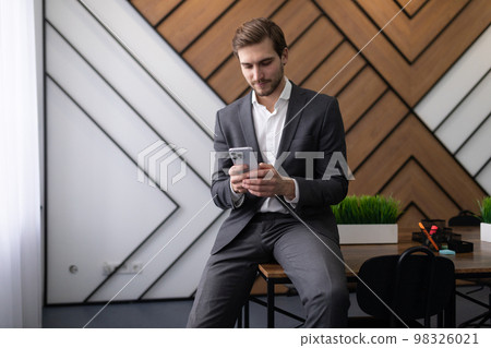 businessman sitting on the desk in the office reading messages on the phone businessman sitting on the desk in the office reading messages on the phone 98326021