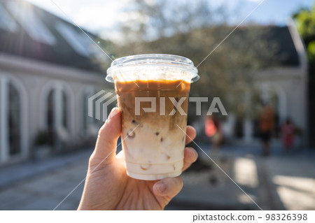 Hand holding iced latte in plastic glass on outside garden at coffee shop 98326398