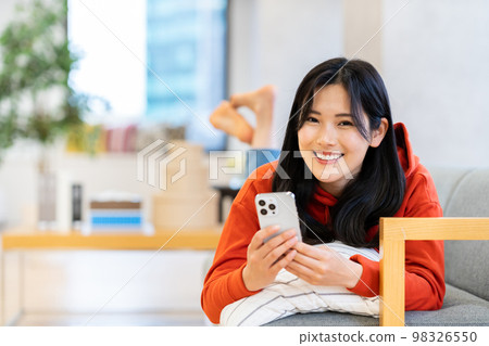 A young woman who operates a smartphone while lying on a sofa. A young woman who operates a smartphone while lying on a sofa. 98326550