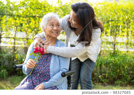 Caregiver daughter hug and help Asian senior or elderly old lady woman holding red rose on wheelchair in park. 98327085