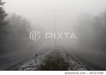 Car traveling on the foggy road with headlights or headlamps on. Low visibility - Dangerous driving of cars in winter in bad weather. Car traveling on the foggy road with headlights or headlamps on. Low visibility - Dangerous driving of cars in winter in bad weather. 98327390