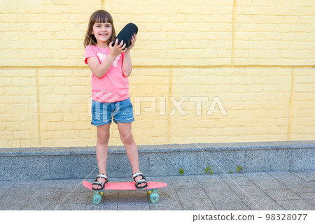 Stylish little girl with a skateboard listens to music on a yellow background 98328077