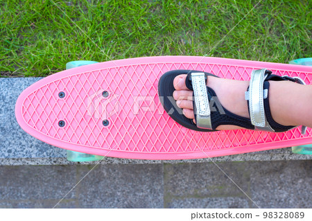 A close up of a child's leg in the sandals standing on a pink plastic skateboard, the top view. Sports lifestyle A close up of a child's leg in the sandals standing on a pink plastic skateboard, the top view. Sports lifestyle 98328089