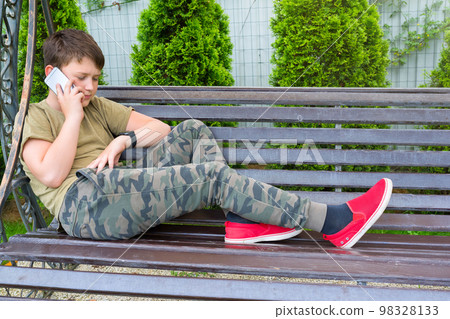 A teenager sitting on the bench, talking over the phone in the open air A teenager sitting on the bench, talking over the phone in the open air 98328133