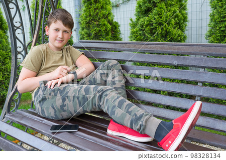 An open portrait of an 11-year-old boy sitting on the bench with a phone and smart watch on his hand An open portrait of an 11-year-old boy sitting on the bench with a phone and smart watch on his hand 98328134