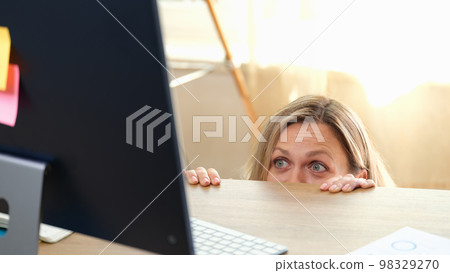 Woman hides behind office desk and looks at computer monitor. 98329270