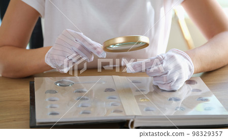 Numismatist in gloves examines collection of coins through magnifying glass. Numismatist in gloves examines collection of coins through magnifying glass. 98329357