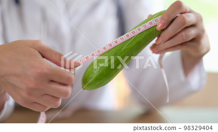 Doctor hands holding long vegetable and measuring tape above table in medical clinic. Doctor hands holding long vegetable and measuring tape above table in medical clinic. 98329404