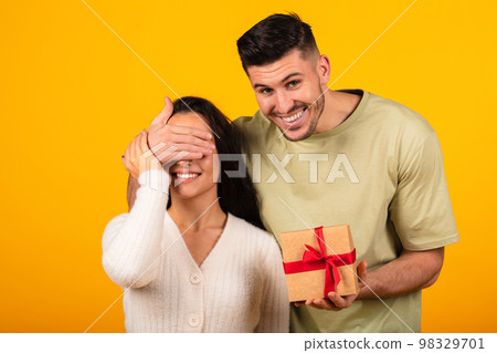 Smiling millennial arab guy closes eyes to woman, gives gift in box, isolated on orange background, studio Smiling millennial arab guy closes eyes to woman, gives gift in box, isolated on orange background, studio 98329701