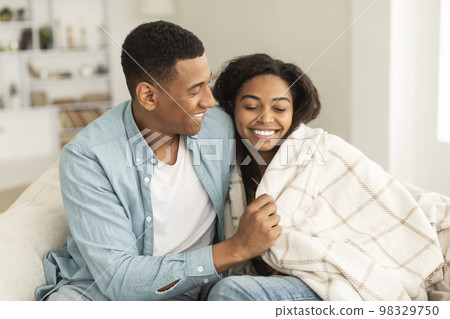 Cheerful young african american guy wraps lady in blanket, couple enjoys warm together, sitting on sofa Cheerful young african american guy wraps lady in blanket, couple enjoys warm together, sitting on sofa 98329750