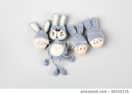 Three Easter eggs with funny faces and gray crocheted hats with bunny ears and a crocheted gray bunny on a gray background. Happy Easter concept. Greeting card. 98330176