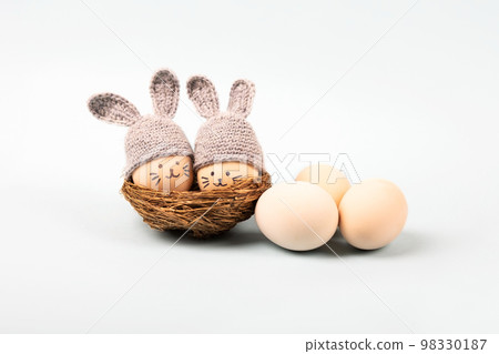 Easter eggs with funny faces and gray crocheted hats with bunny ears in a decorative nest of sisal on a gray background. Happy Easter concept. Greeting card. 98330187
