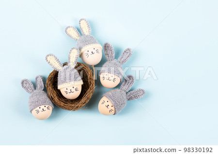 Five Easter eggs with funny faces and gray crocheted hats with bunny ears in a decorative nest of sisal on a blue background. Happy Easter concept. Greeting card. 98330192