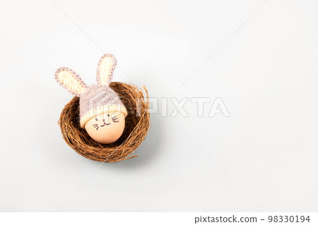 Easter egg with funny face and gray crocheted hat with bunny ears in a decorative nest of sisal on a gray background. Happy Easter concept. Greeting card. Easter egg with funny face and gray crocheted hat with bunny ears in a decorative nest of sisal on a gray background. Happy Easter concept. Greeting card. 98330194