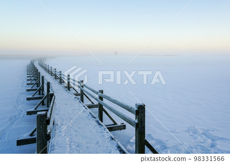 Wooden boardwalk on the frozen lake leading to the bird watching tower, winter sunset landscape Wooden boardwalk on the frozen lake leading to the bird watching tower, winter sunset landscape 98331566