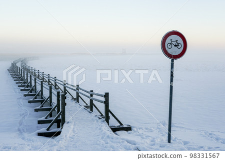 No bicycles road sign next to a snowy wooden boardwalk on a frozen lake, misty winter landscape No bicycles road sign next to a snowy wooden boardwalk on a frozen lake, misty winter landscape 98331567