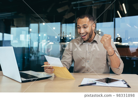 Successful businessman received notification letter with good news, man inside office opens and reads mail envelope with message celebrate victory success, hispanic man holds hand up triumph gesture. 98331898