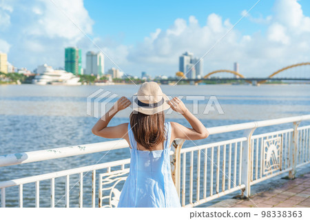Woman Traveler with blue dress visiting in Da Nang city. Tourist sightseeing the river view with Dragon bridge. Landmark and popular for tourist attraction. Vietnam and Southeast Asia travel concept 98338363