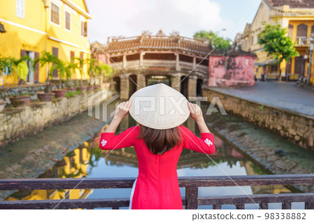 Woman traveler wearing Ao Dai Vietnamese dress sightseeing at Japanese covered bridge in Hoi An town, Vietnam. landmark and popular for tourist attractions. Vietnam and Southeast Asia travel concept 98338882