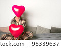 A woman sits in bed with inflatable balloons of red color. Valentine's Day, the concept of the holiday. Front view. 98339637