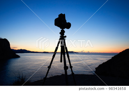 Photo camera silhouette on tripod at rocky beach 98341658