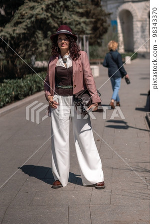 Woman park city. Stylish woman in a hat walks in a park in the city. Dressed in white corset trousers and a pink jacket with a bag in her hands. Woman park city. Stylish woman in a hat walks in a park in the city. Dressed in white corset trousers and a pink jacket with a bag in her hands. 98343370