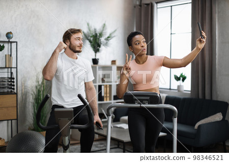 African woman running on treadmill and fit caucasian man cycling bike and making selfie 98346521
