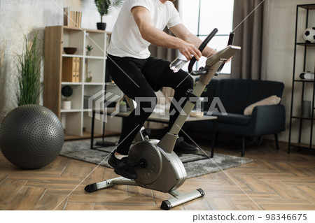 Cropped view of young male with water bottle wearing sportswear using exercise bike. 98346675