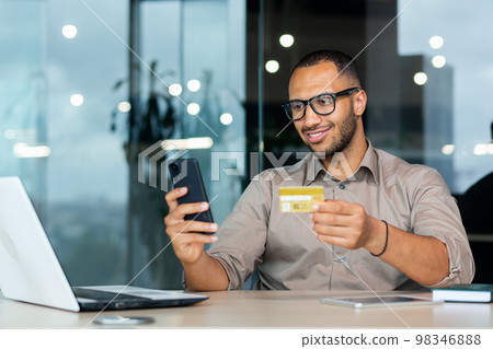 Successful hispanic businessman inside office, man in shirt smiling and happy holding bank credit card and phone, making money transfer and online shopping. Successful hispanic businessman inside office, man in shirt smiling and happy holding bank credit card and phone, making money transfer and online shopping. 98346888