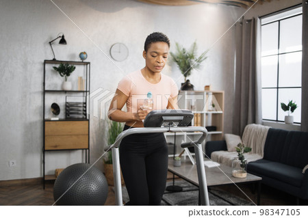 African woman in sports clothes and headphones holding bottle of water doing cardio training 98347105