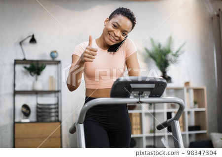 Front view of african sporty woman in pink t-shirt working out in treadmill while having phone call Front view of african sporty woman in pink t-shirt working out in treadmill while having phone call 98347304