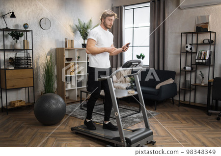 Caucasian sportsman having online video class training on smartphone using treadmill 98348349