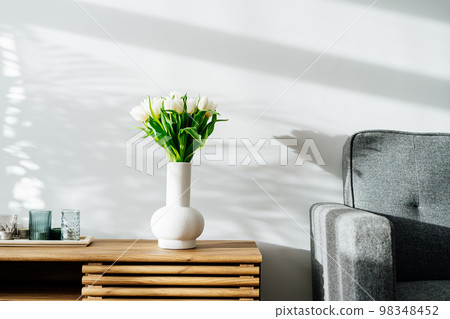 Scandinavian home interior with spring bouquet of white tulip flowers in ceramic vase, tray with candles standing on a wooden cabinet. Minimalist design with gray sofa and white wall. Springtime. 98348452