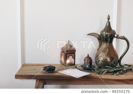 Ramadan Kareem still life. Ornamental burning lantern, cup and golden arabic dallah coffee pot. Green olive tree branches and date fruit on old wooden table. White wallbackground. Muslim Iftar dinner. Ramadan Kareem still life. Ornamental burning lantern, cup and golden arabic dallah coffee pot. Green olive tree branches and date fruit on old wooden table. White wallbackground. Muslim Iftar dinner. 98350170