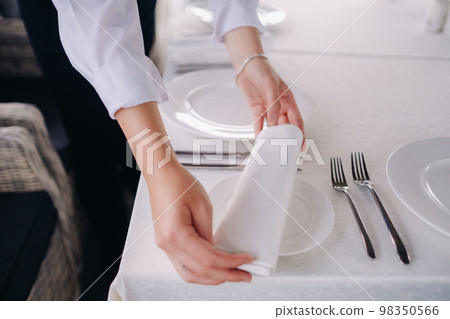 The waiter puts a white napkin on the plate. Beautiful table setting for a holiday without food The waiter puts a white napkin on the plate. Beautiful table setting for a holiday without food 98350566