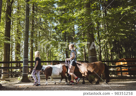 Side view of two young girls walking their ponies around in the forest - Horse ranch 98351258