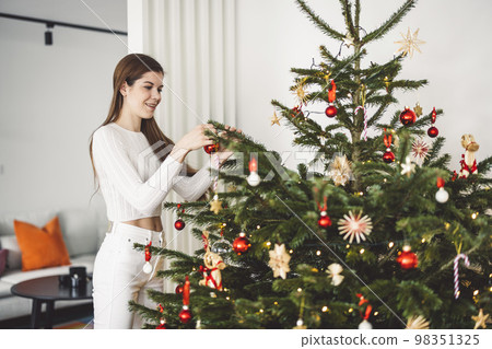 Young woman dressed in white outfit decorating the Christmas tree  98351325