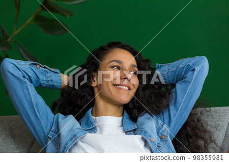 Beautiful hispanic woman daydreaming sitting on couch at home and looking out window, happy woman with hands behind head close up. 98355981