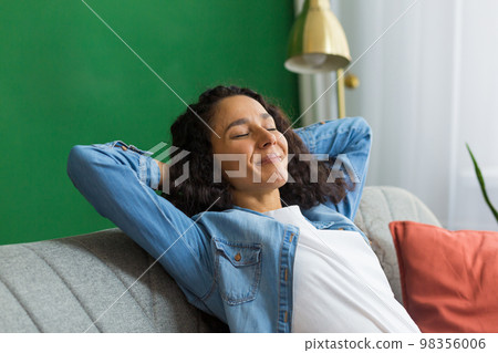 Beautiful hispanic woman at home close up with closed eyes relaxing sitting on couch, woman with hands behind head smiling in living room. Beautiful hispanic woman at home close up with closed eyes relaxing sitting on couch, woman with hands behind head smiling in living room. 98356006