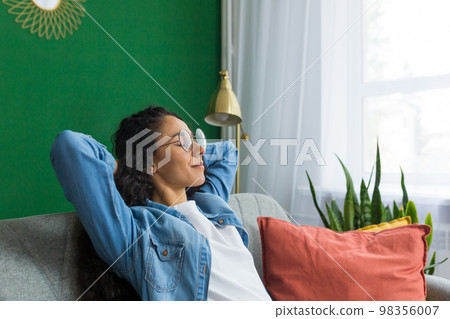 Beautiful hispanic woman at home close up with closed eyes relaxing sitting on couch, woman with hands behind head smiling in living room. 98356007