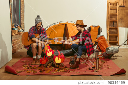 Warming hands over fake fire. Two men imagining, recreating camping activity indoors with necessary equipment. Concept of quarantine, travelling, imagination 98356469